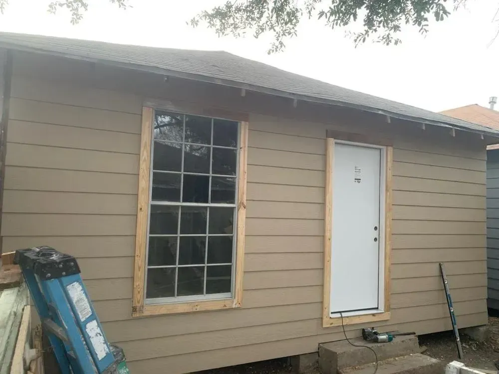 Tan-sided building with a window and a door, both framed in wood. A ladder rests on the ground.