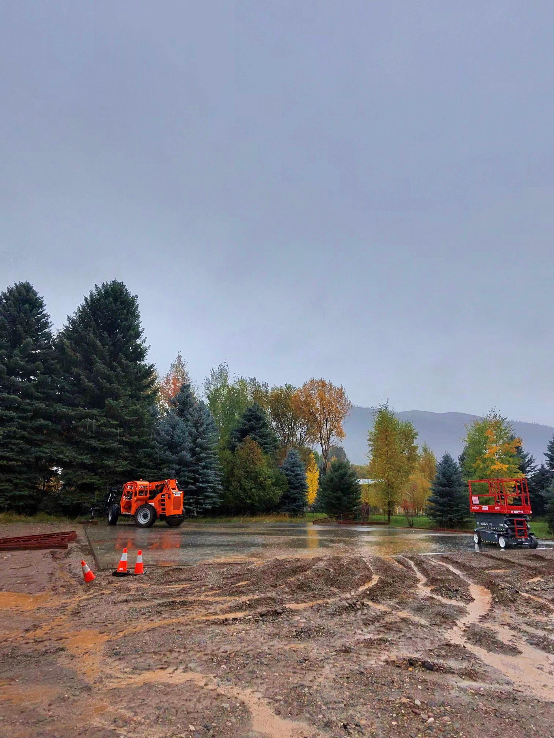 Muddy construction site with orange equipment, surrounded by trees under a cloudy sky.