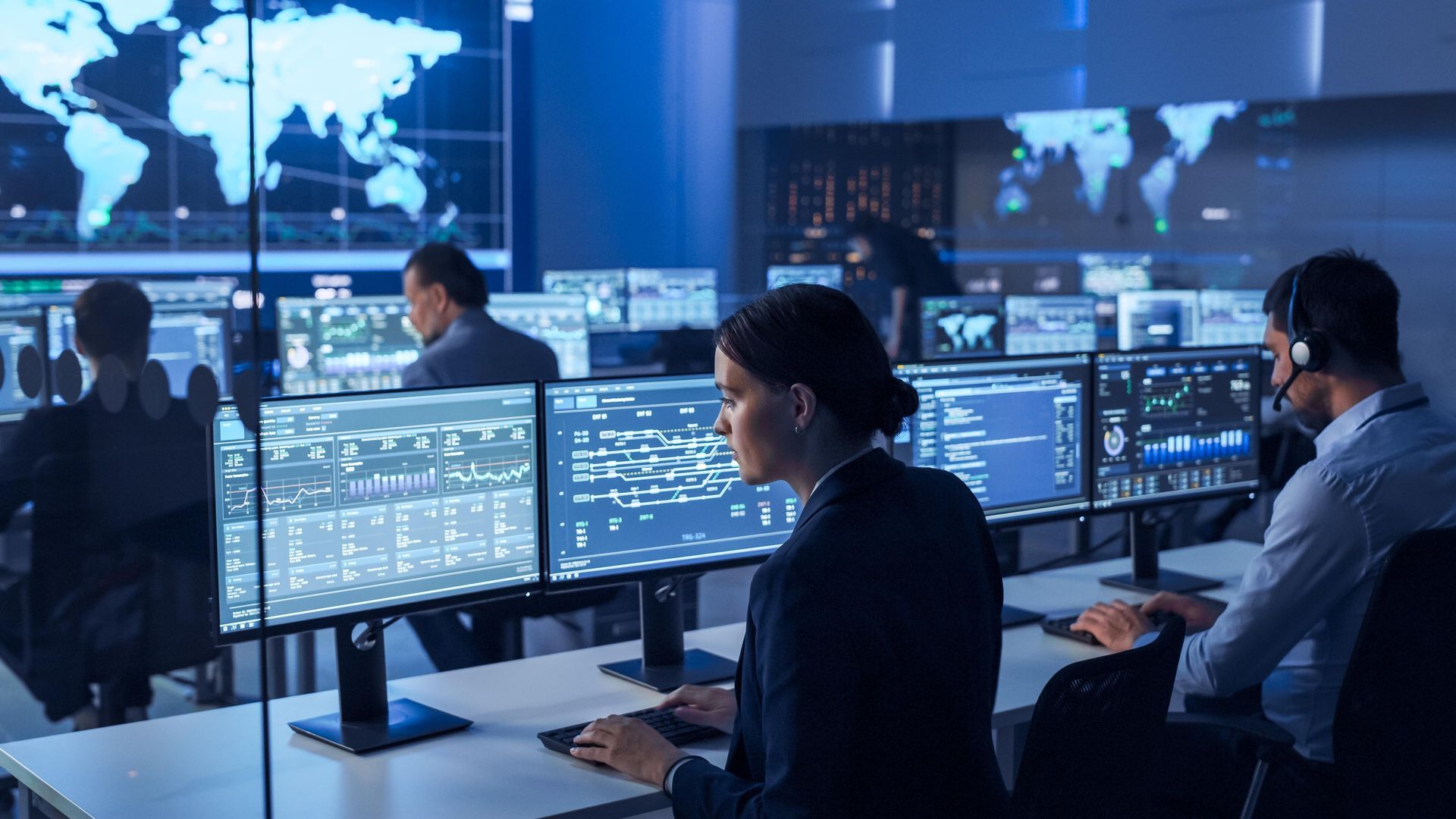 People working in a high-tech control room with multiple computer screens displaying data and world maps. Dim lighting.