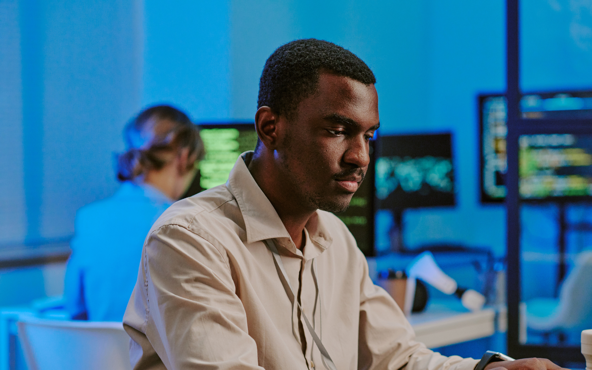 Man working on a computer in a tech-focused office with blue lighting. Others work on computers.