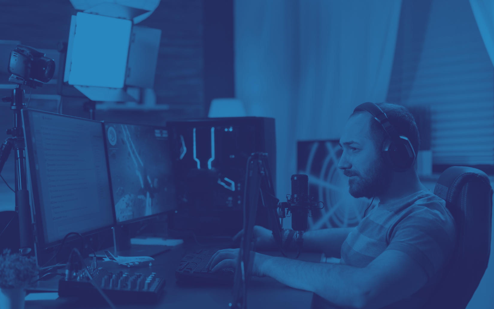 Man wearing headphones in a blue-tinted studio, working at a computer with a microphone, lights, and monitors.