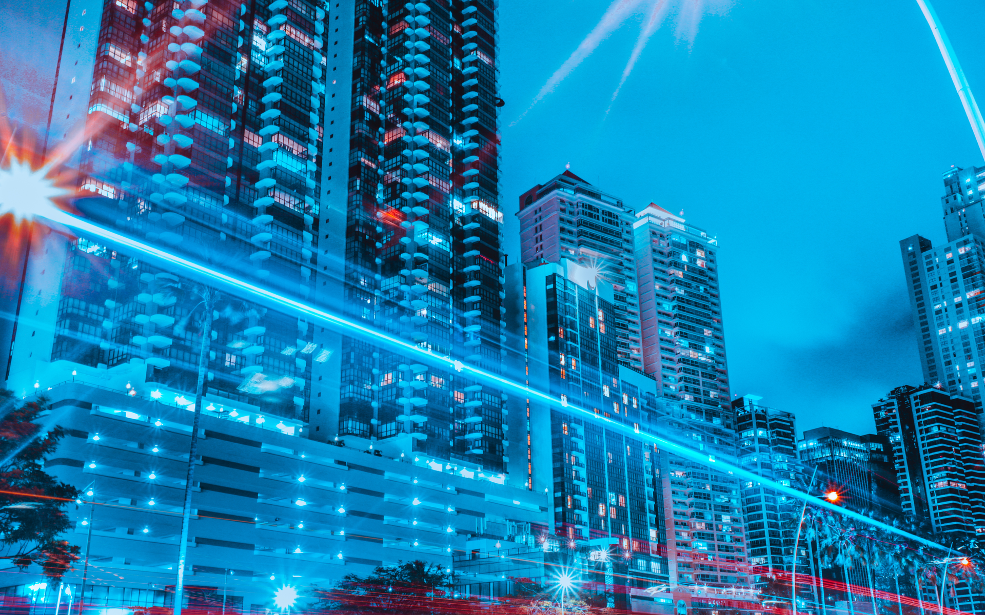 Blue-toned cityscape at dusk with tall buildings, illuminated by lights and streaks of light trails.