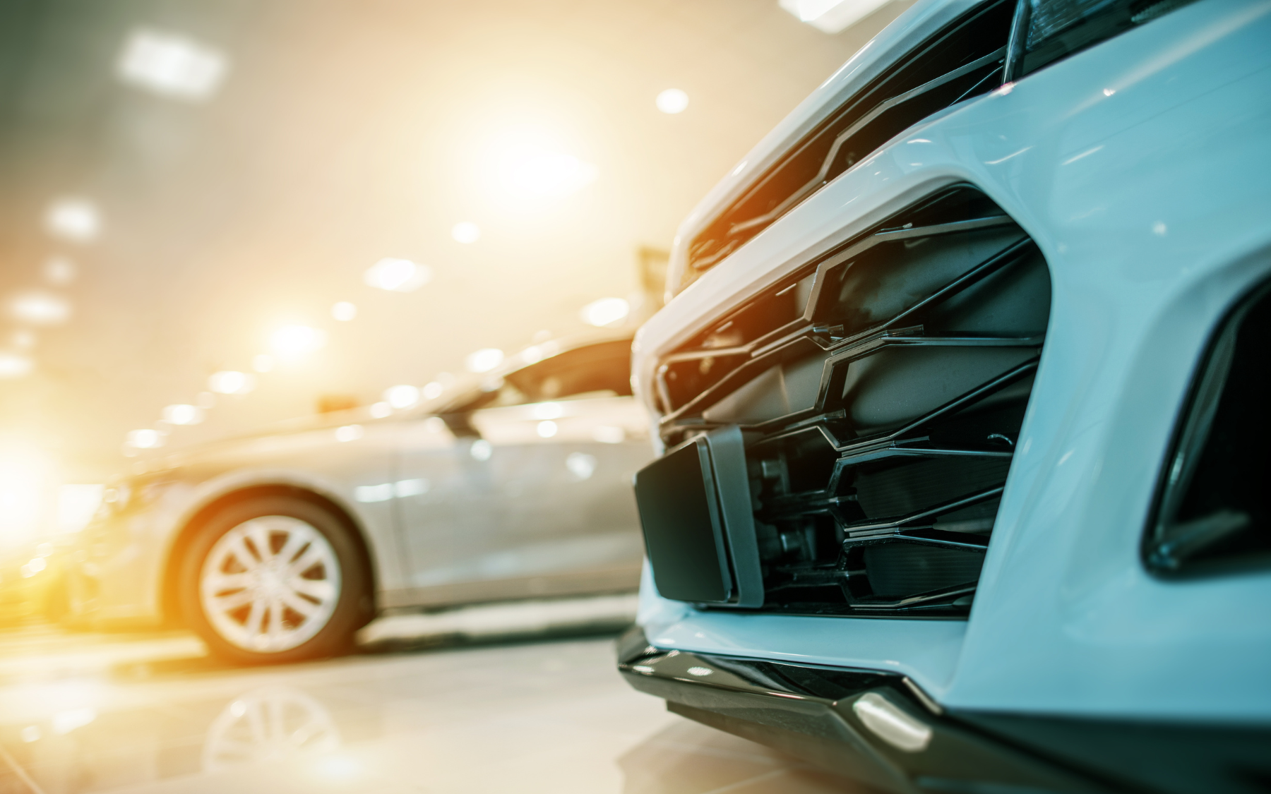Close-up of a white car's front, with a blurred silver car in the background, inside a brightly lit showroom.