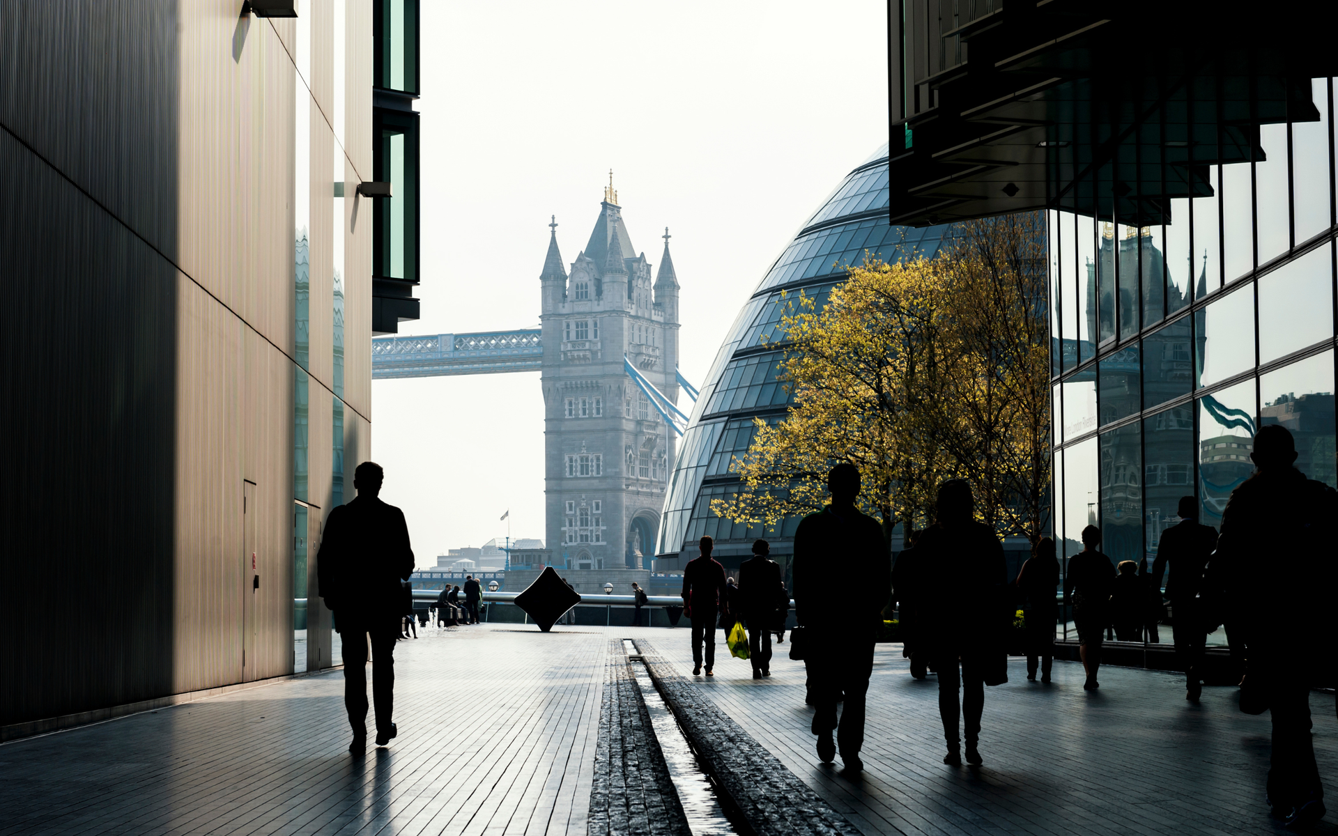 People walk toward Tower Bridge in London, a modern glass building and the City Hall dome are in the background.