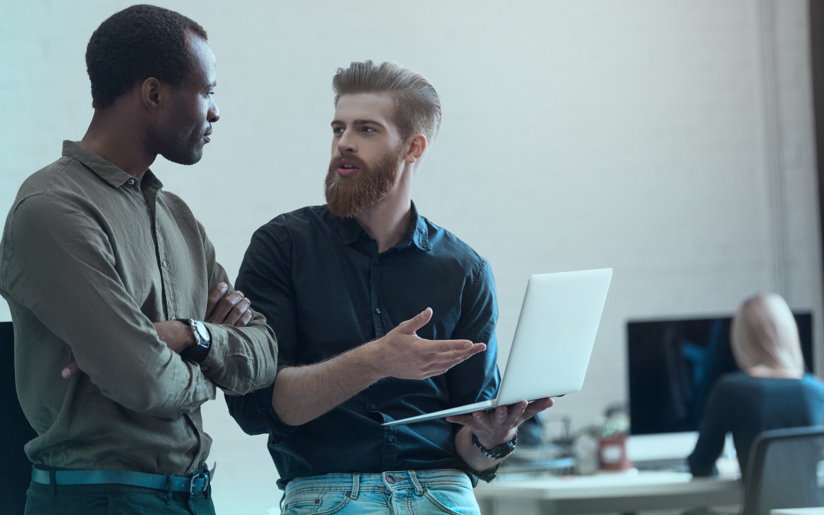 Two men are standing next to each other in an office looking at a laptop.