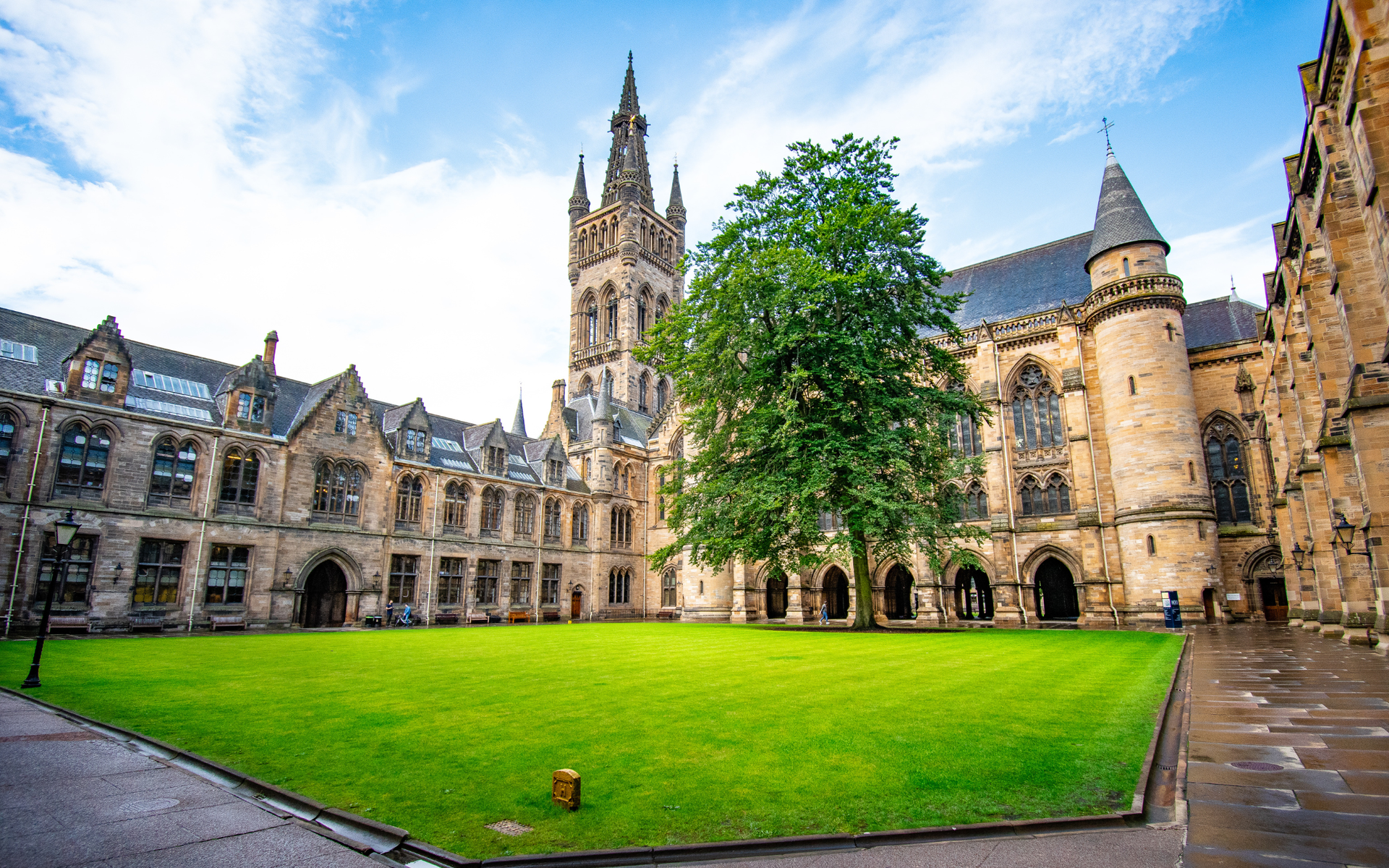 University of Glasgow quad with lush green lawn, stone buildings, and a tall tower under a partly cloudy sky.