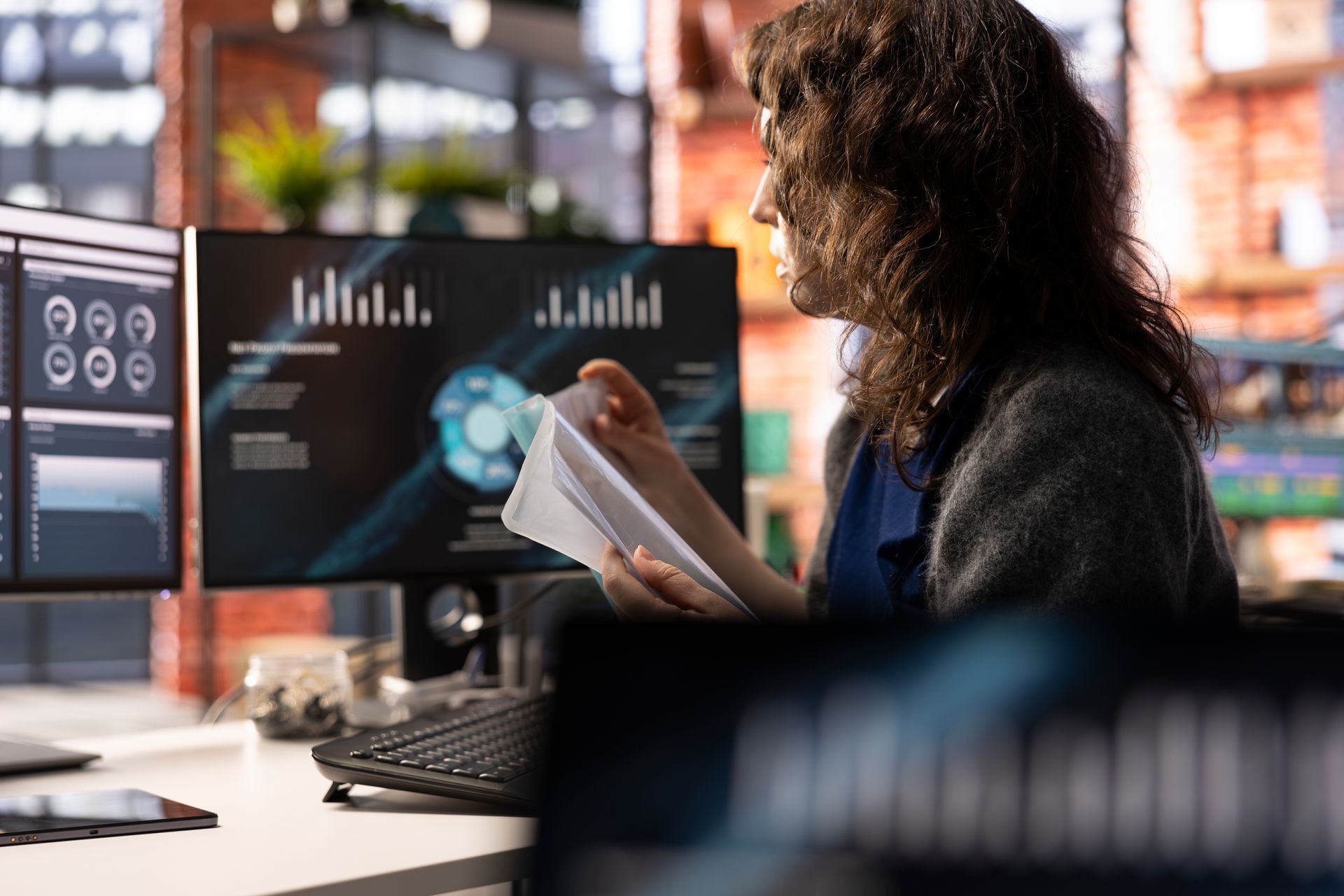 A woman is sitting at a desk in front of a computer holding a piece of paper.