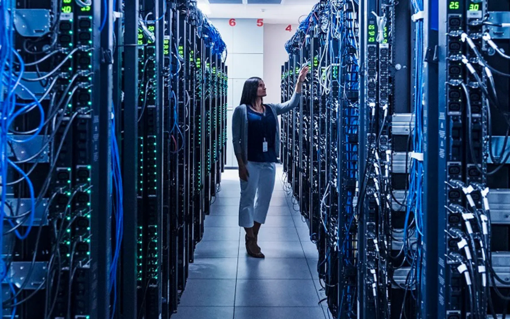 Woman in a server room checks equipment, surrounded by rows of blinking servers and cables.