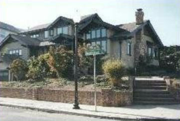 A large, multi-story house with a brick and stone facade, shrubs, and a street sign.