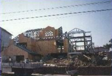House under construction, wood and metal framing visible. Debris and workers visible.