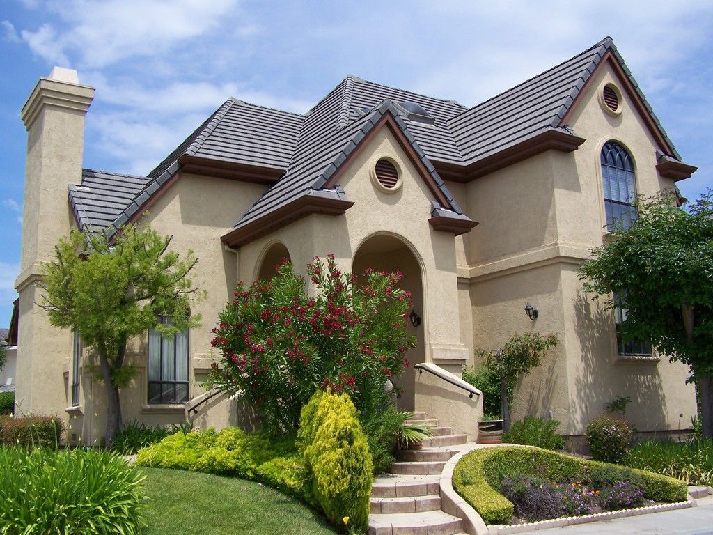 Tan house with dark roof and trim, arched windows, and landscaping against a blue sky.