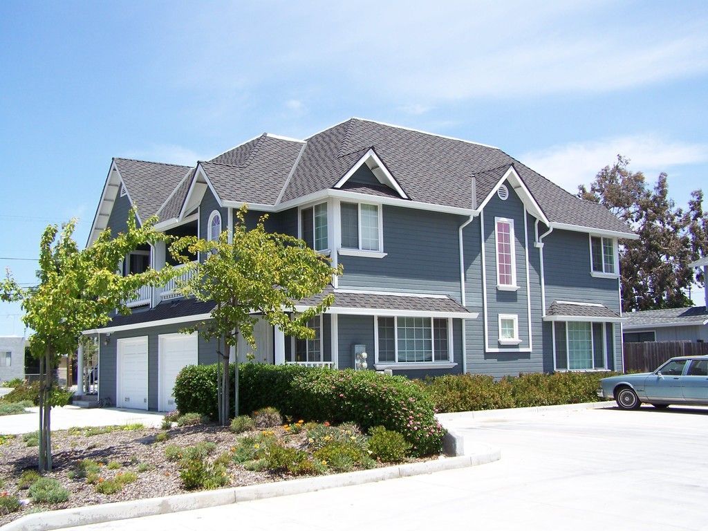 Two-story blue house with white trim, three-car garage, and landscaped yard on a sunny day.