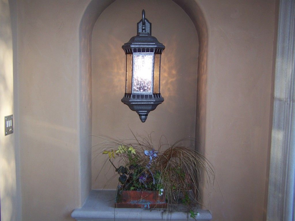 Lantern inside a recessed wall niche, above a small planter with dried grasses and flowers. Light beige walls.
