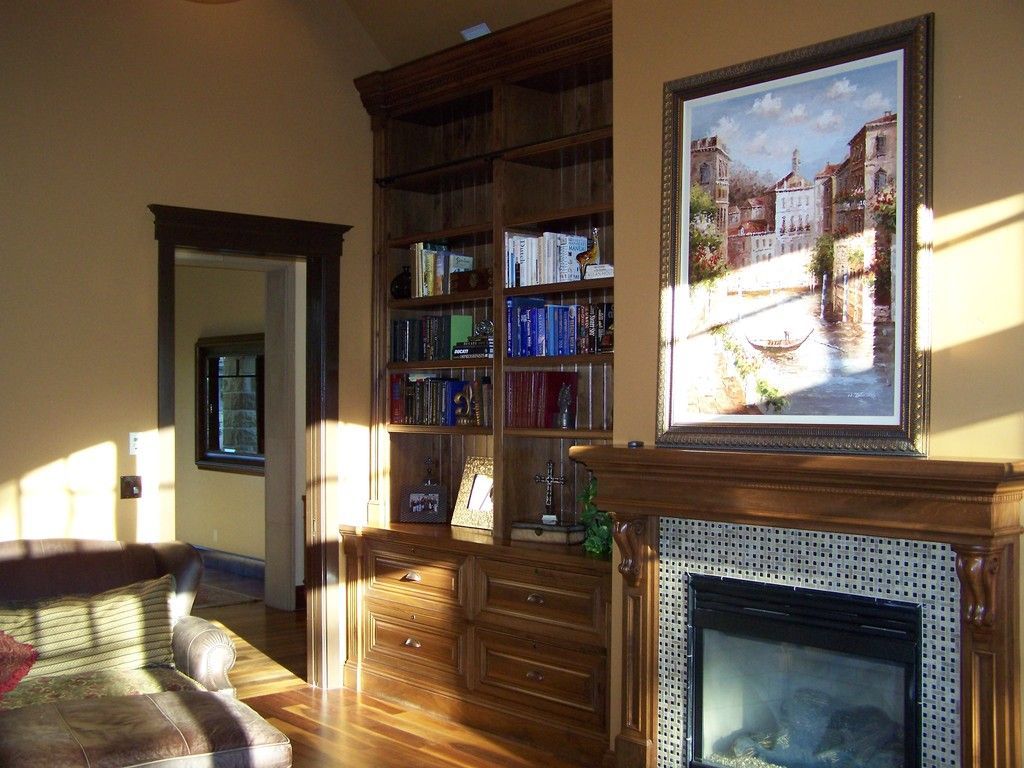 Living room with built-in bookshelves, fireplace, and painting. Warm lighting, wooden floors, tan walls, and a sofa.