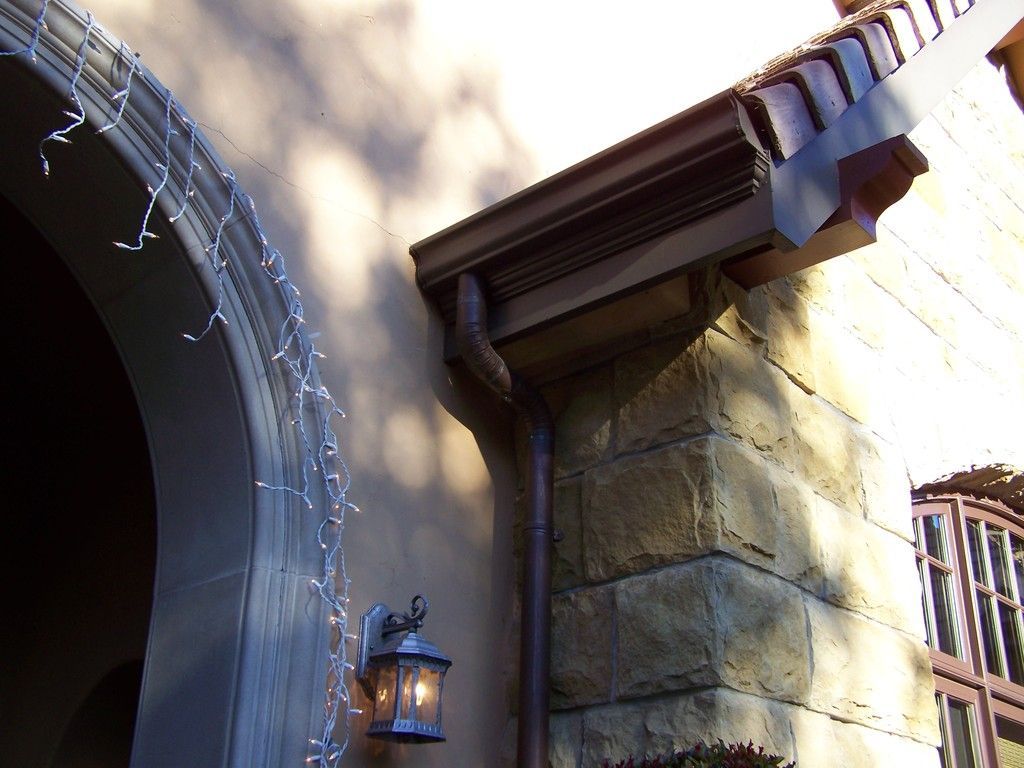 Close-up of a house roof edge with clay tiles, brown gutters, and a decorative brown support.