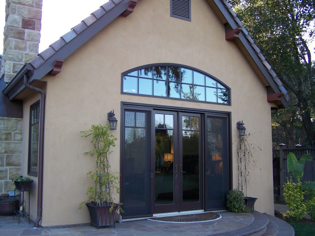 Beige stucco building with dark brown arched doors and windows, potted plants, and stone chimney.