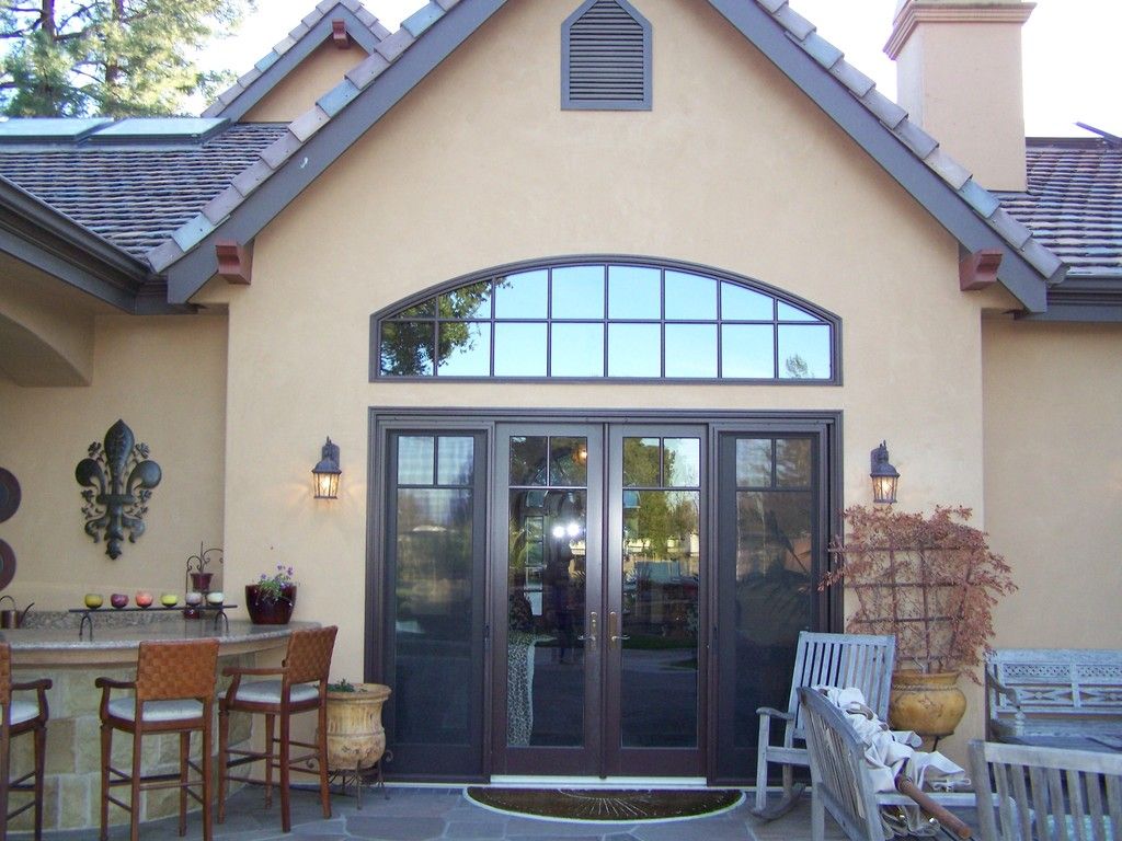 Tan stucco two-story house with arched doorway, windows, and brown roof. Green lawn and shrubs in front.