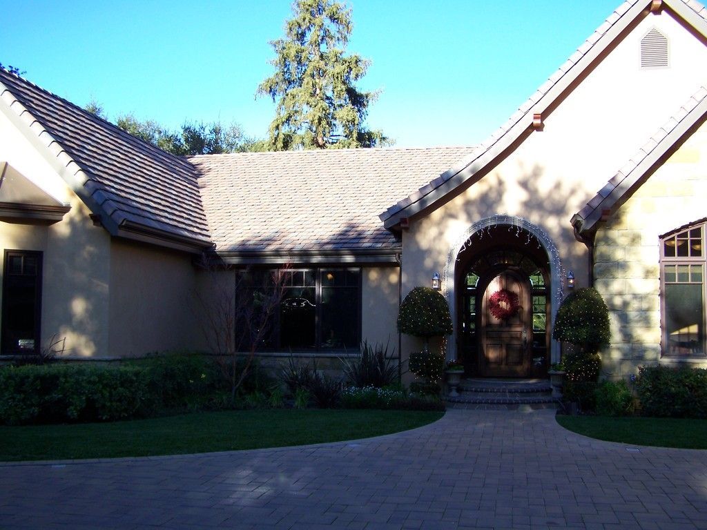 Beige stucco house with tile roof and arched entryway. Paved driveway, shrubs, and trees.