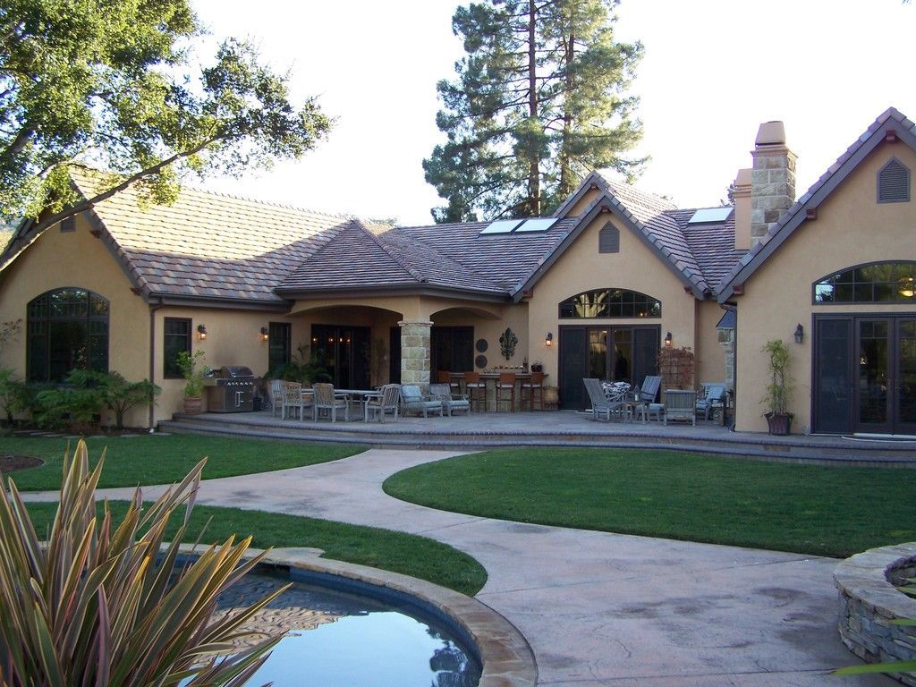 Backyard view of a tan house with a tile roof, patio furniture, and a small pool on a sunny day.