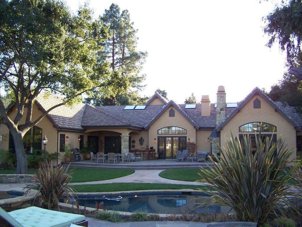 Backyard view of a large house with a pool and patio. Tan stucco exterior, clay tile roof, and tall trees.