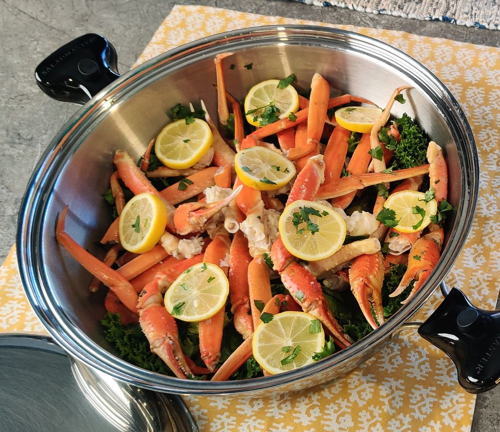 Crab legs with lemon slices and parsley in a silver bowl on a yellow patterned surface.