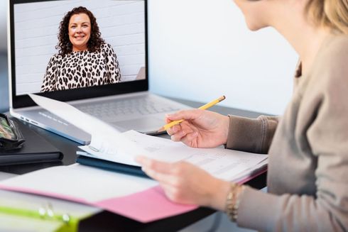 Woman on a video call with another woman, taking notes. The setting is a home office, with papers and a laptop visible.