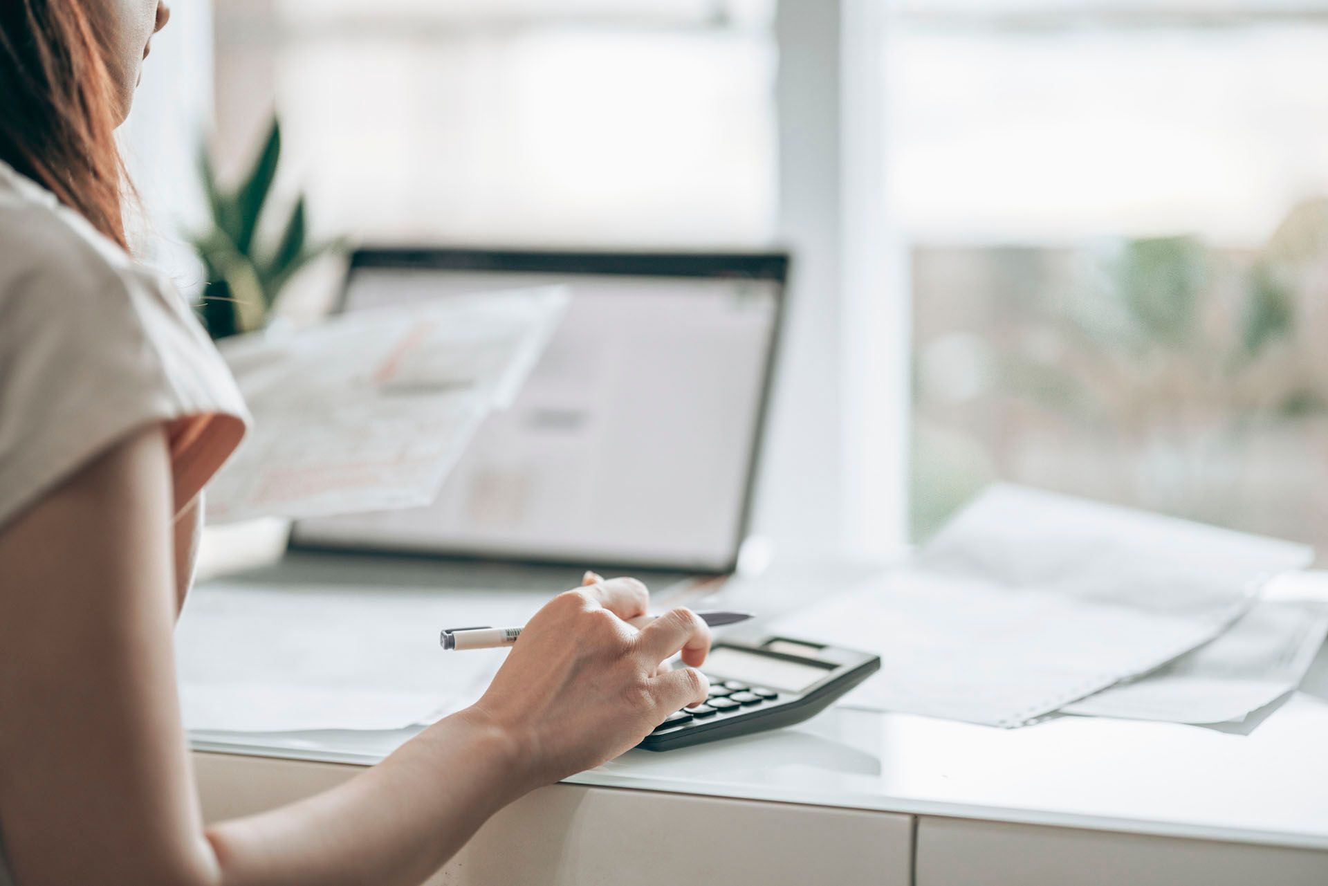 Woman calculating finances with a calculator, laptop and papers on a white desk near a window.