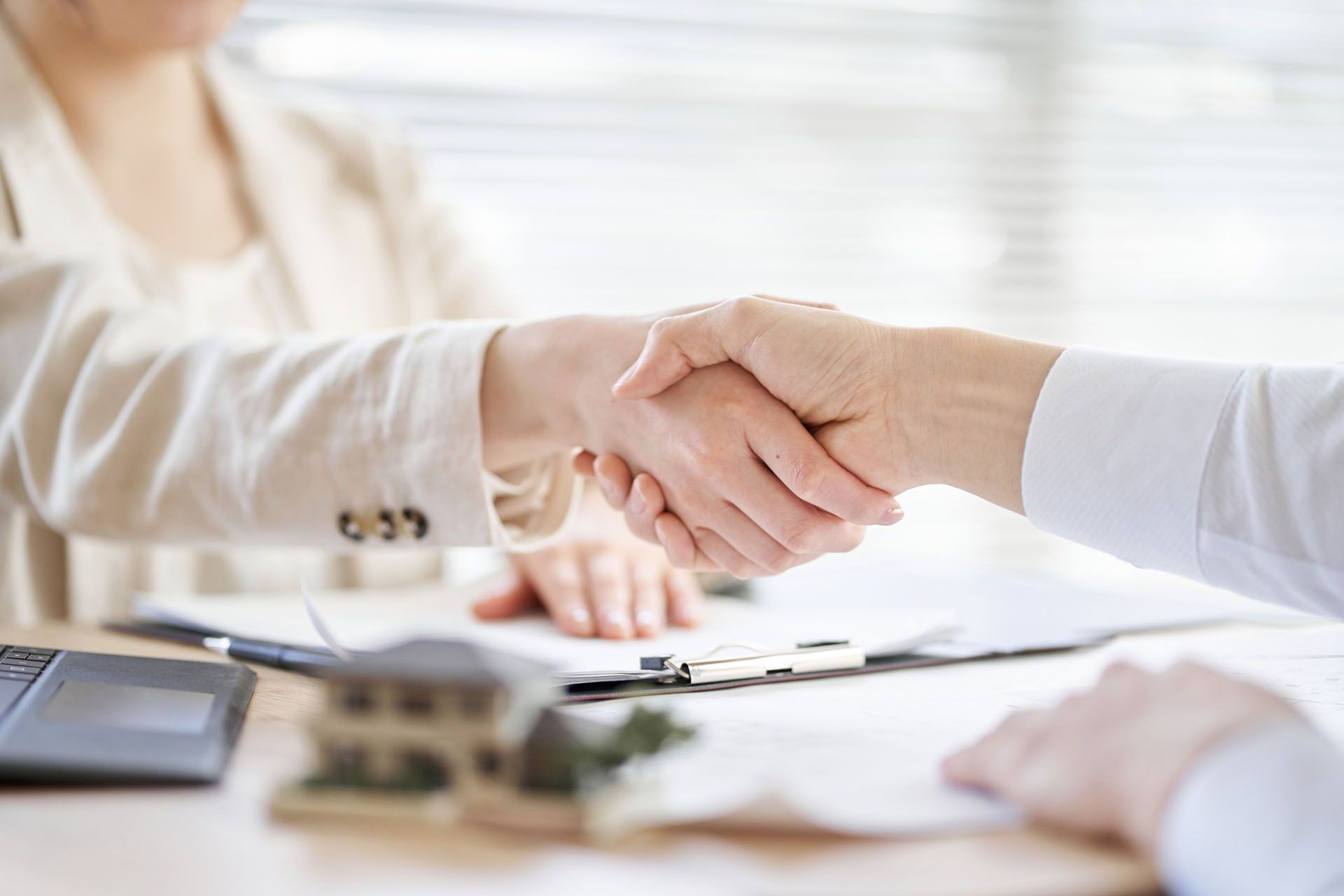 Two people shaking hands over a document, possibly closing a real estate deal. A model house and a laptop are on the table.