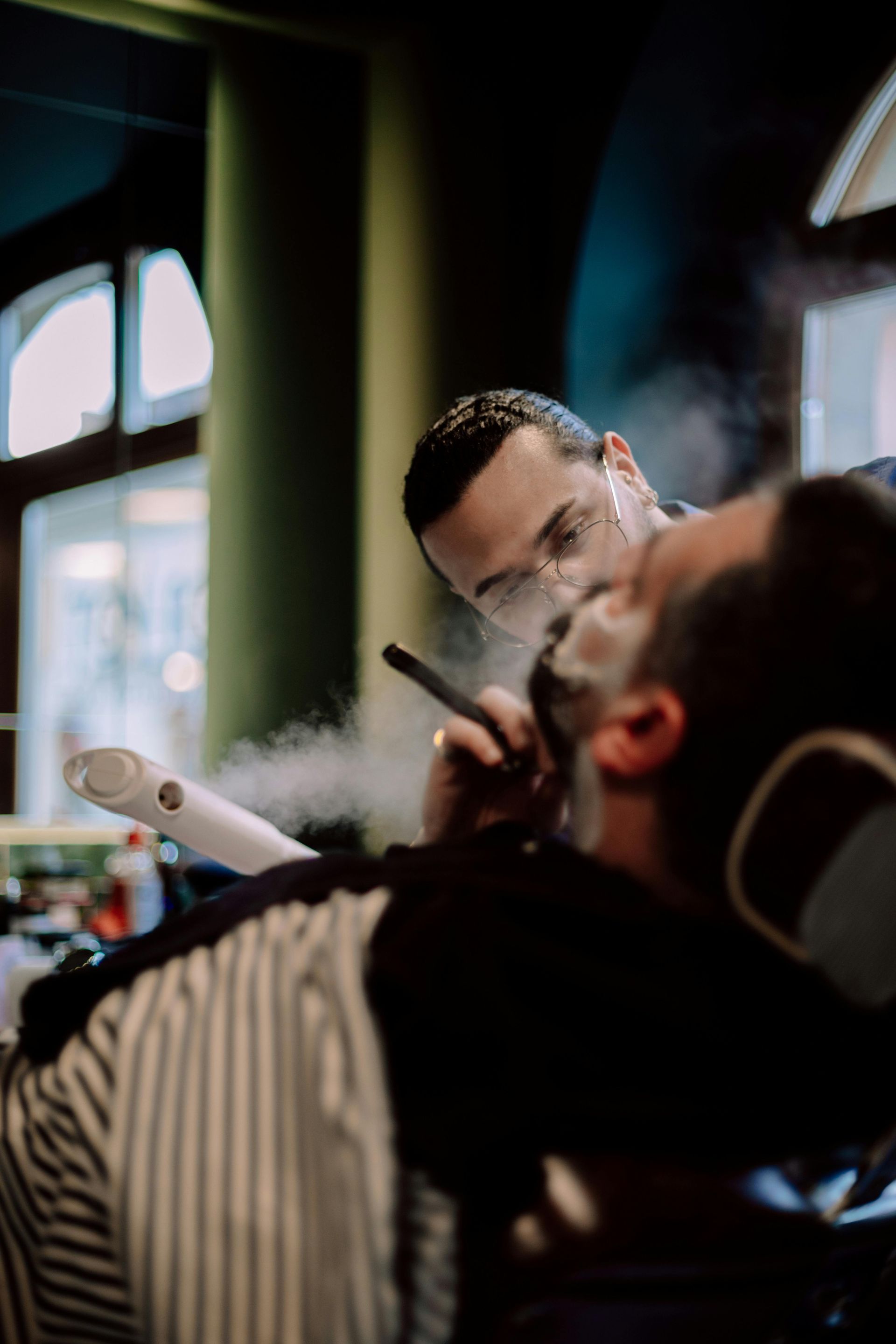 Barber steaming client's face with white vapor, shaving the client's beard in a barbershop.