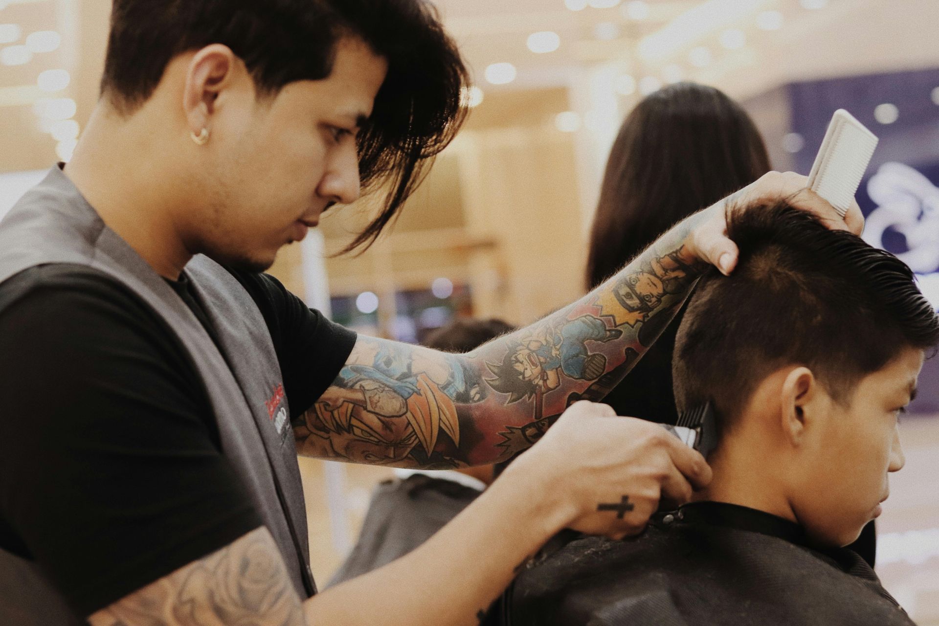 Barber giving a haircut with clippers; tattoos visible on his arm. Child in chair, indoor setting.