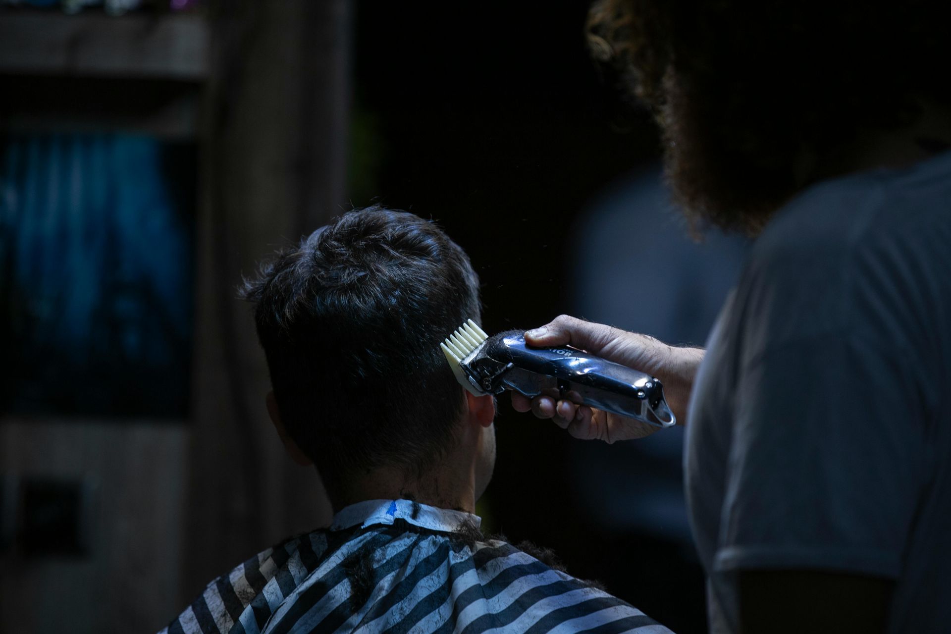 Person getting a haircut with electric clippers in a dimly lit barbershop.