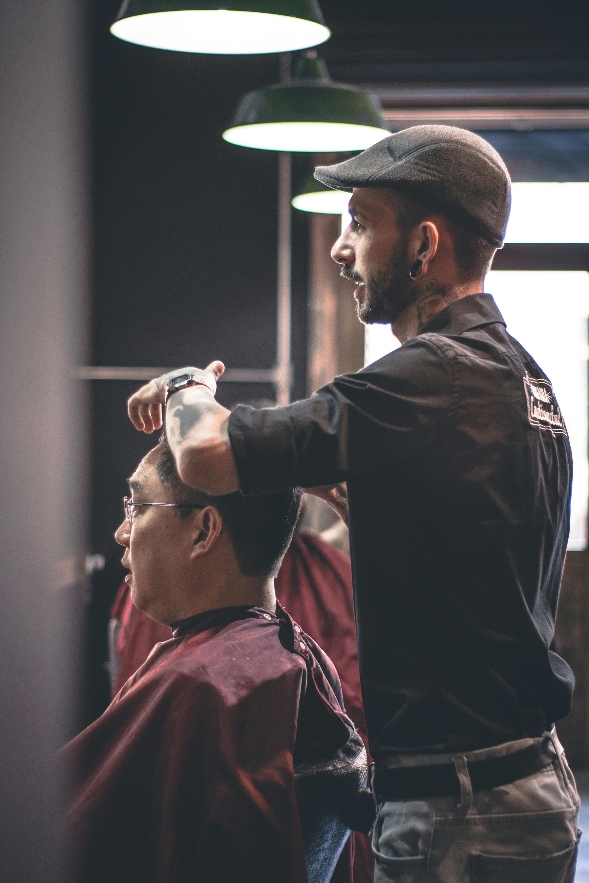 Barber cutting a client's hair in a barber shop. The barber wears a cap and has tattoos, client in a red cape.