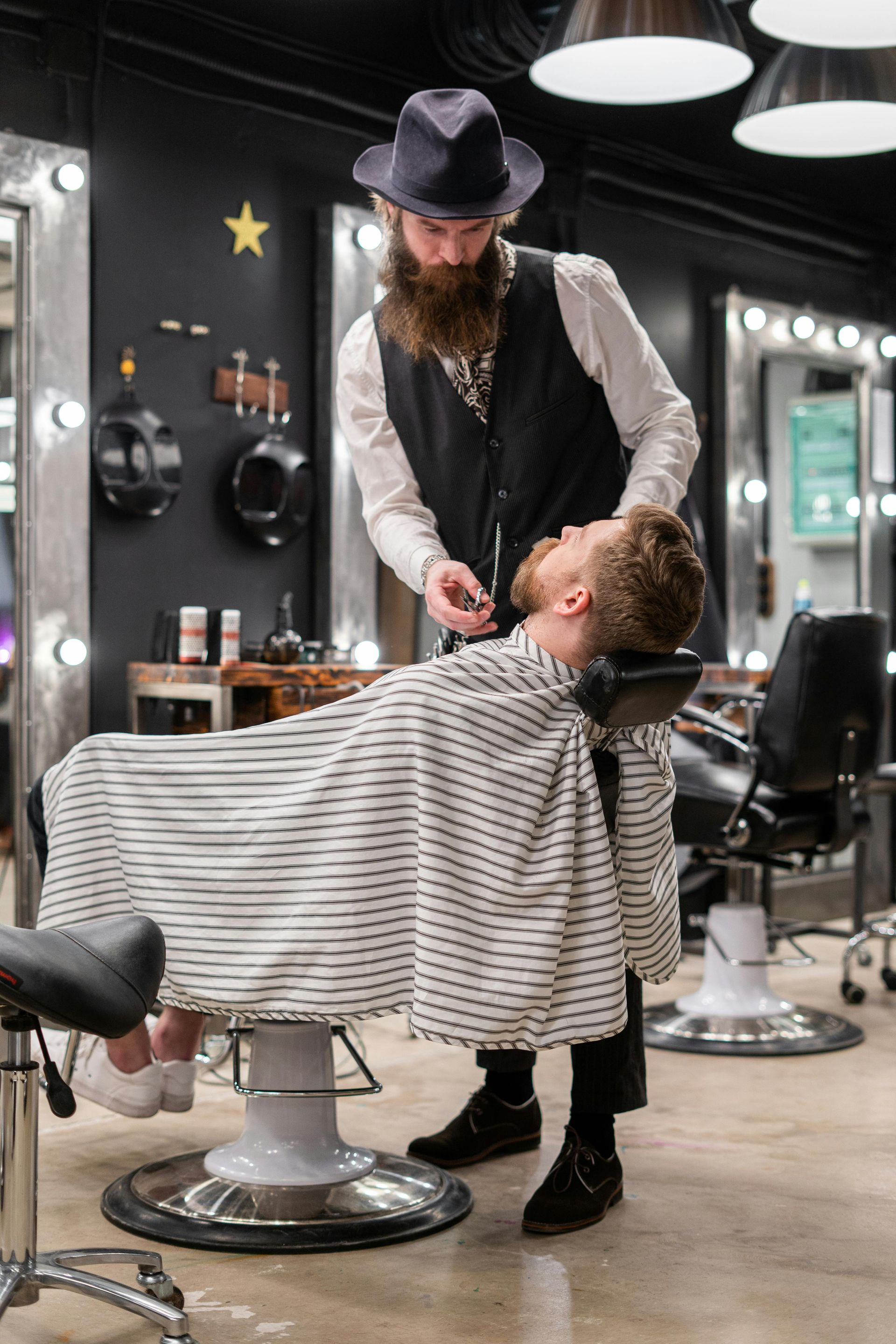 Barber trimming a customer's beard in a stylish salon. The customer is in a chair, draped in a striped cape.