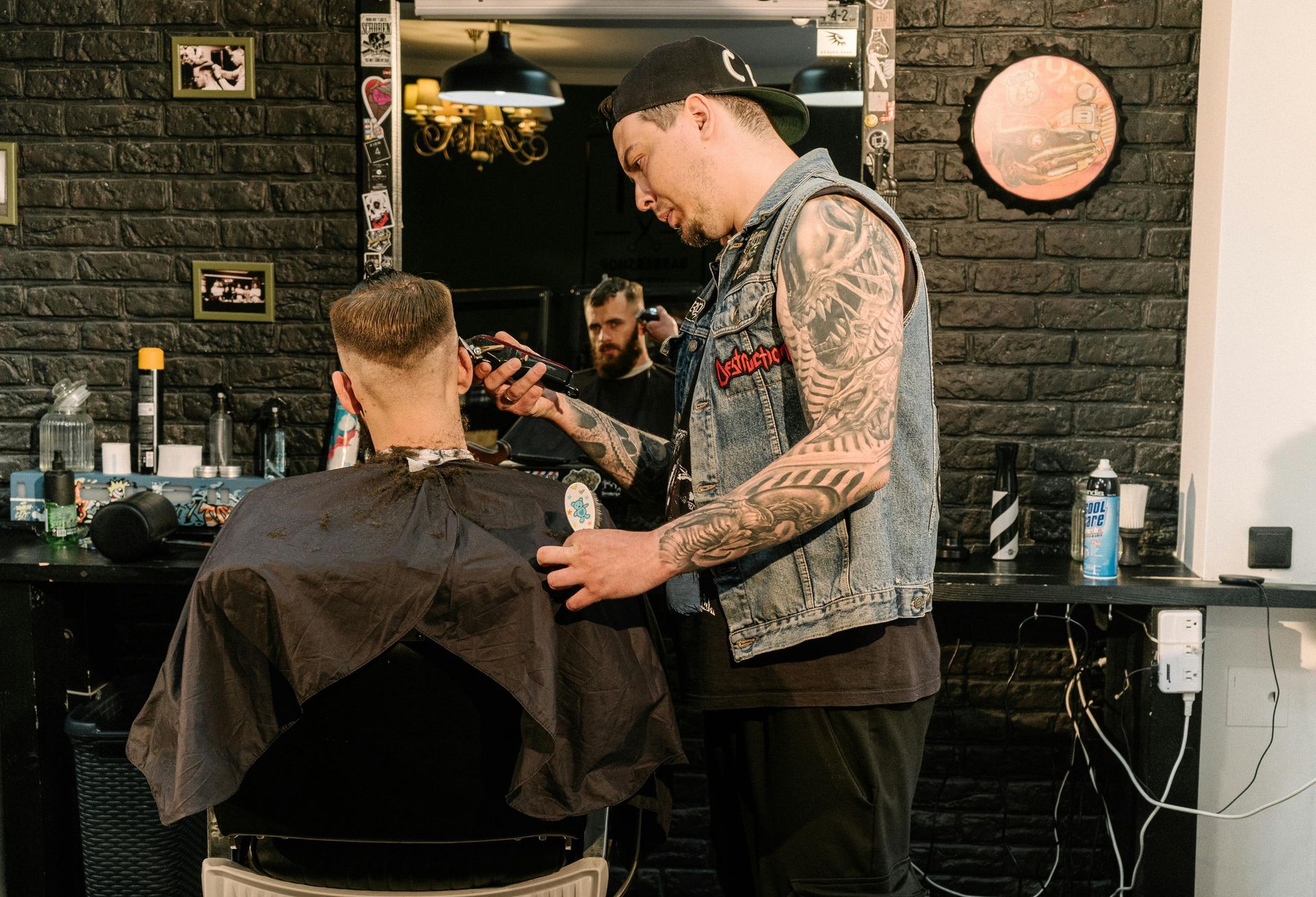 Barber giving a client a haircut in a shop with a black brick wall. The barber has tattoos and the client is in a chair.