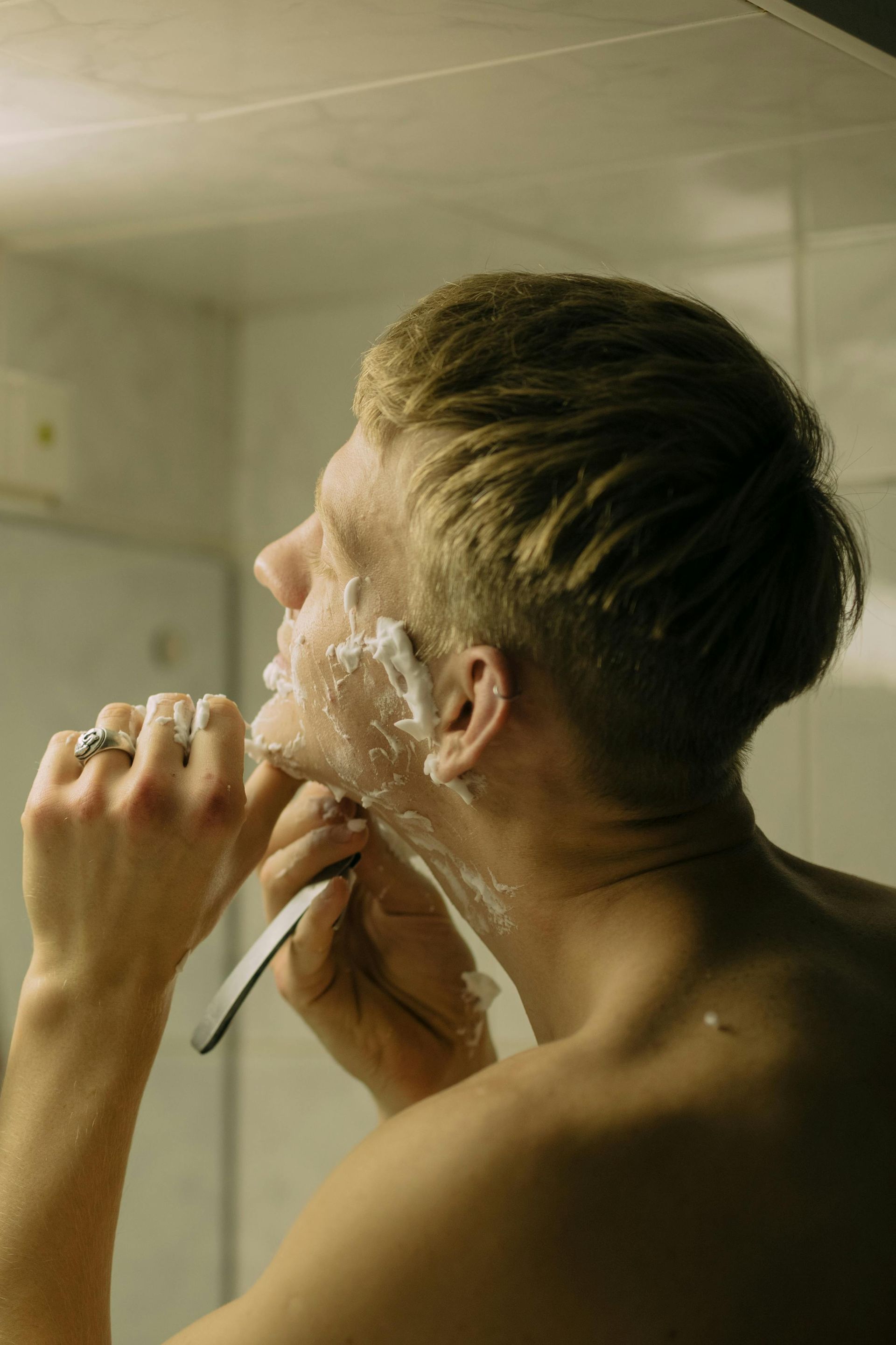 Man shaves face with a straight razor in a bathroom, cream on his skin, looking up at mirror.