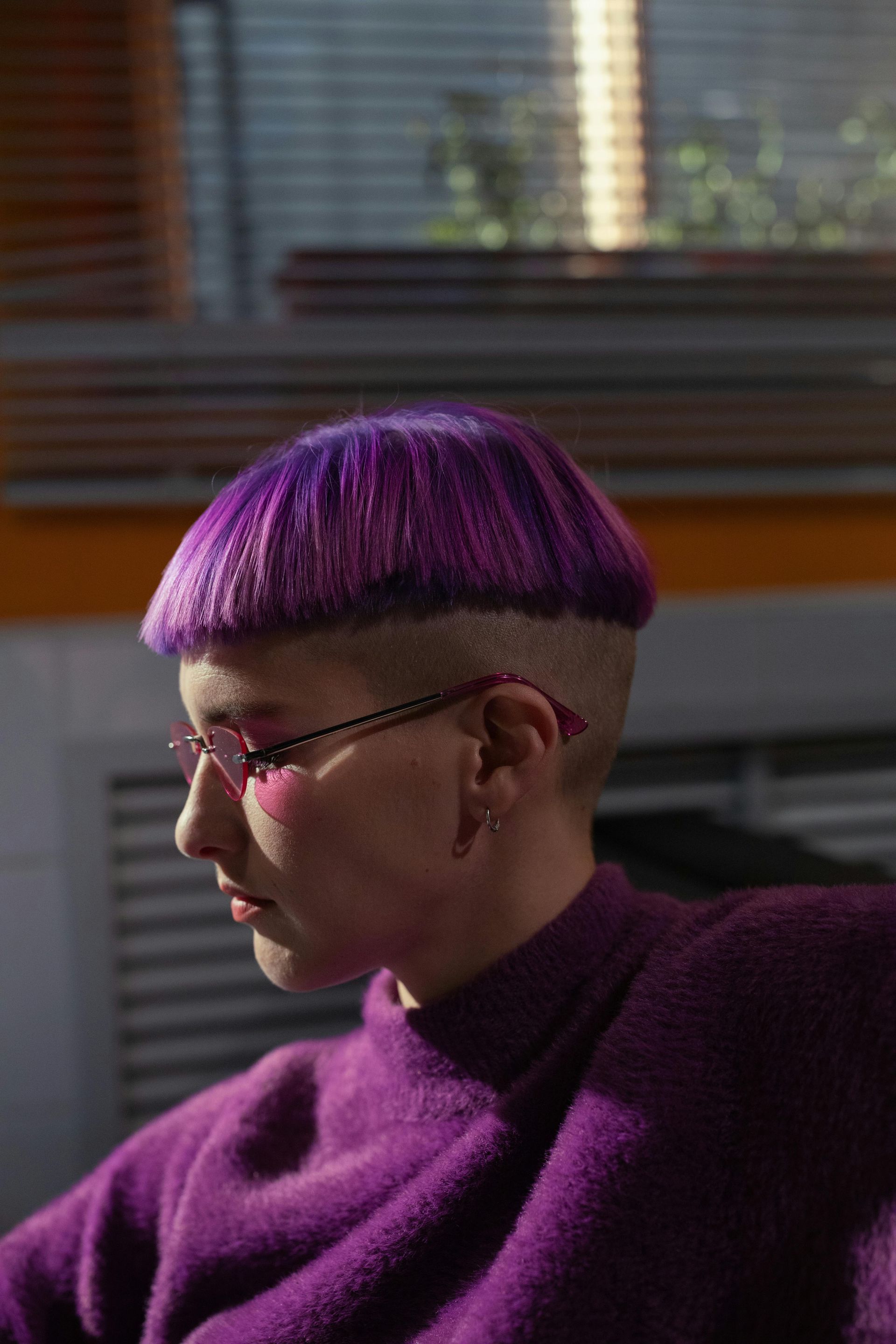 Person with purple bowl cut and glasses, wearing a purple sweater, side profile, indoors.
