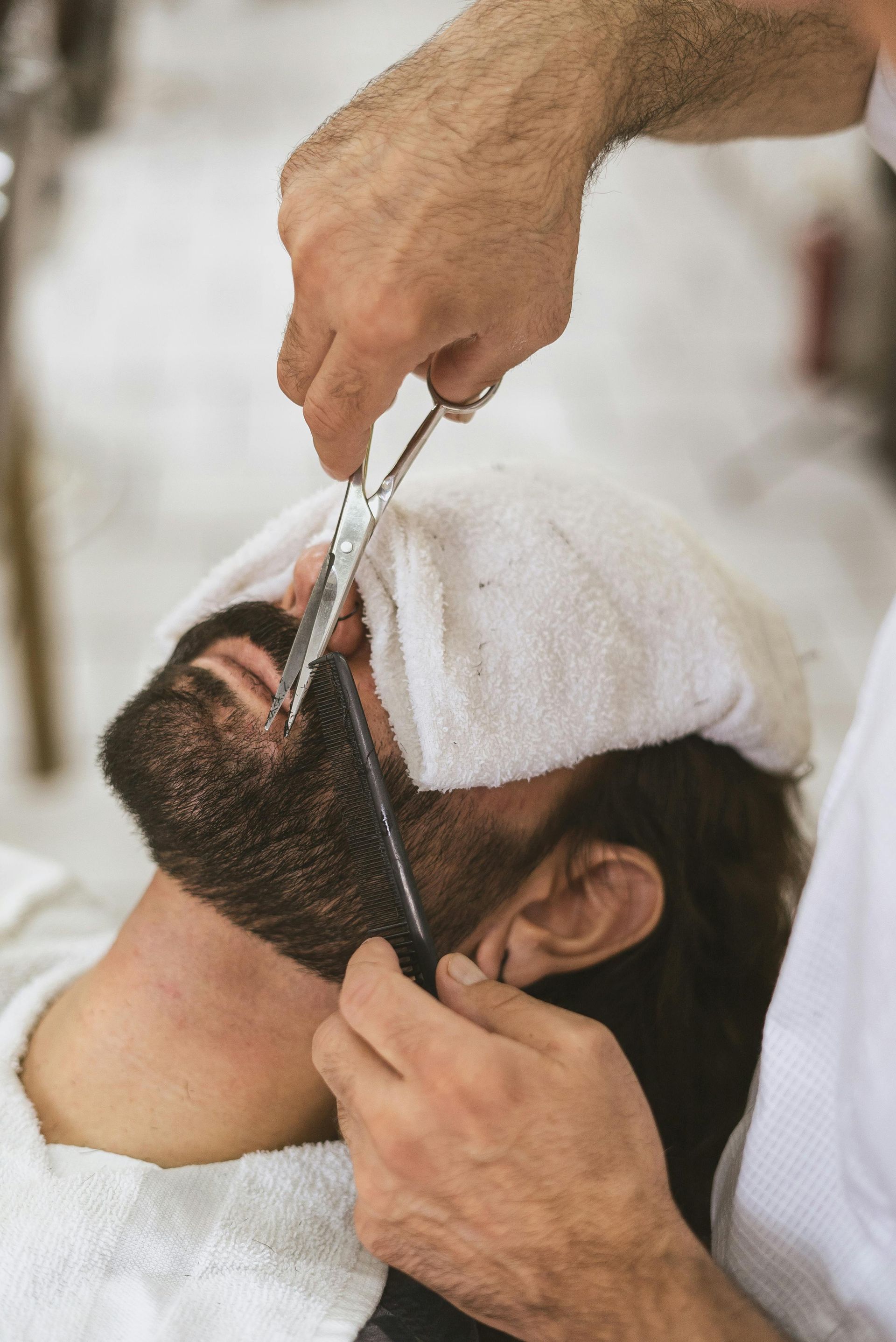 Barber trimming a man's beard with scissors and comb, towel on face.