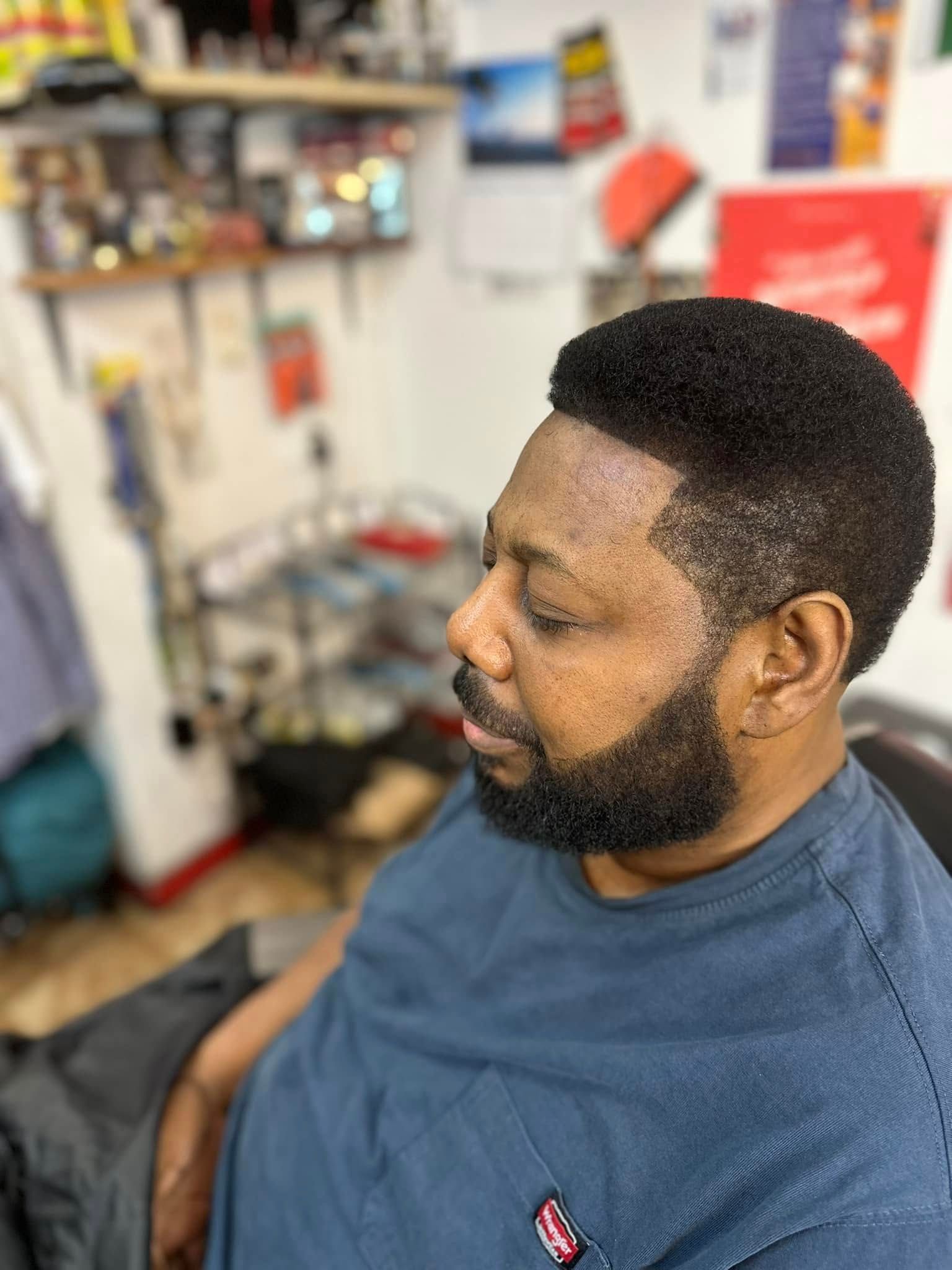 A person with a short, faded haircut and a groomed beard sits in a barbershop, wearing a blue shirt.