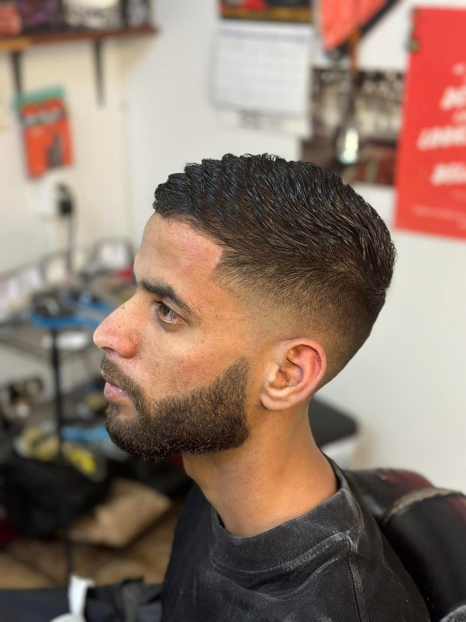 A person with a short, curly textured haircut and a well-groomed beard, seen in profile in a barbershop setting.
