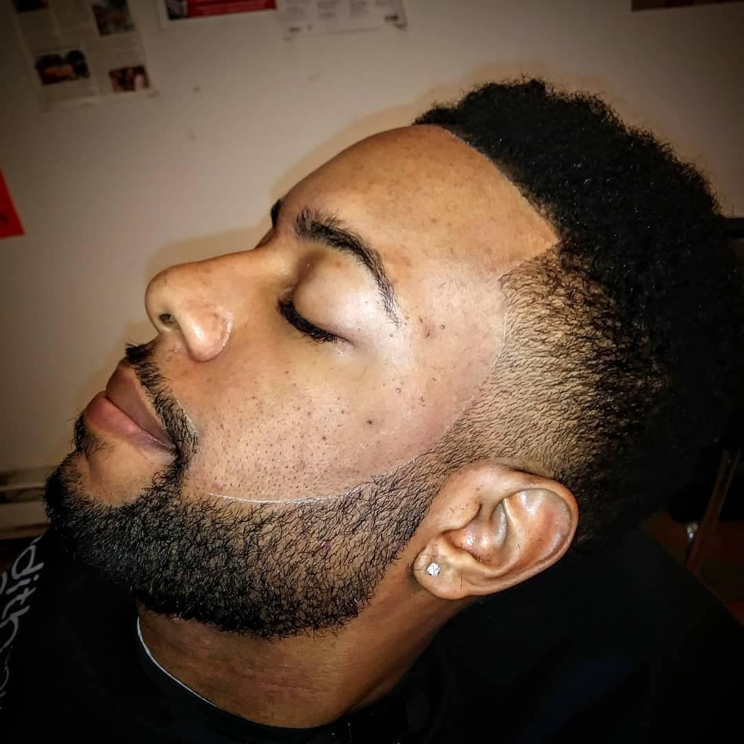 A side-profile shot of a person with a precision-edged beard and a faded haircut, eyes closed, in an indoor setting.