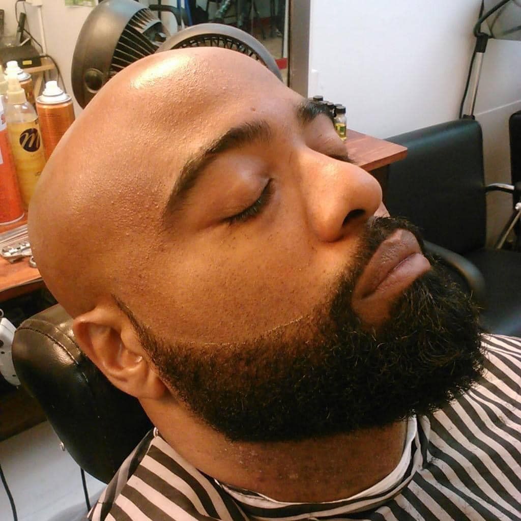 A person with a shaved head and a well-groomed, dark beard sits in a salon chair wearing a striped barber cape.