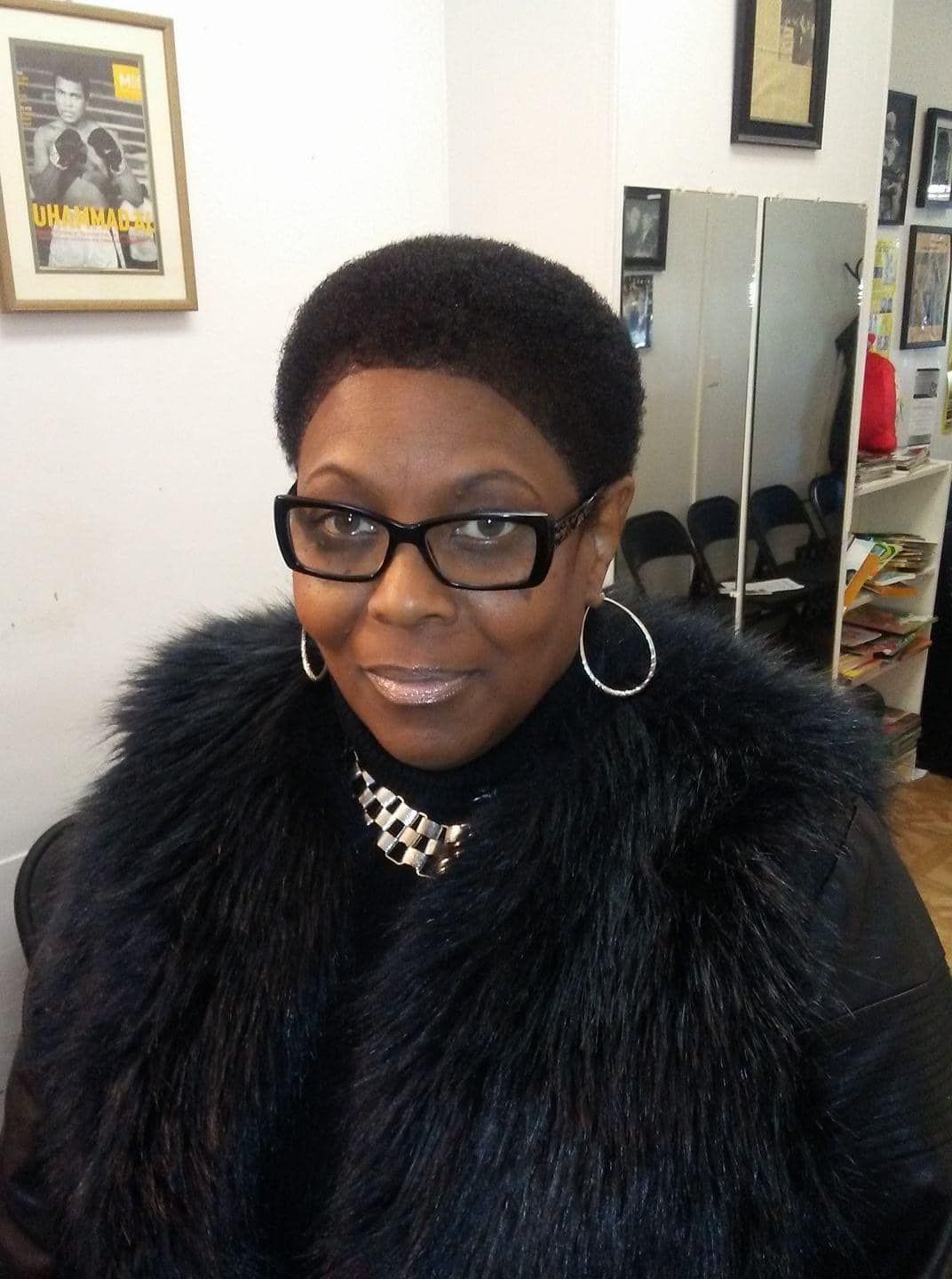 A person with short, dark hair wearing black-rimmed glasses, a black fur-textured jacket, and hoop earrings.