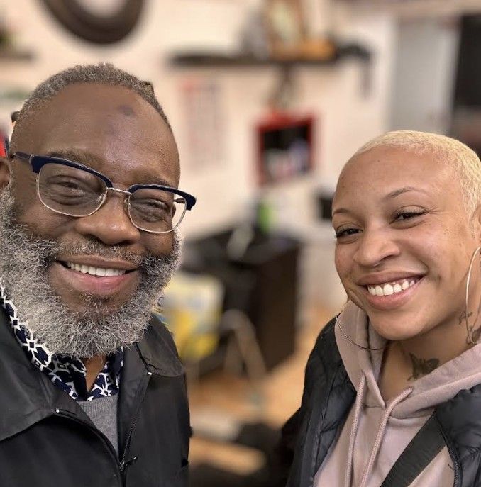 Two people smiling for a selfie in an indoor setting, wearing casual jackets and sweaters.