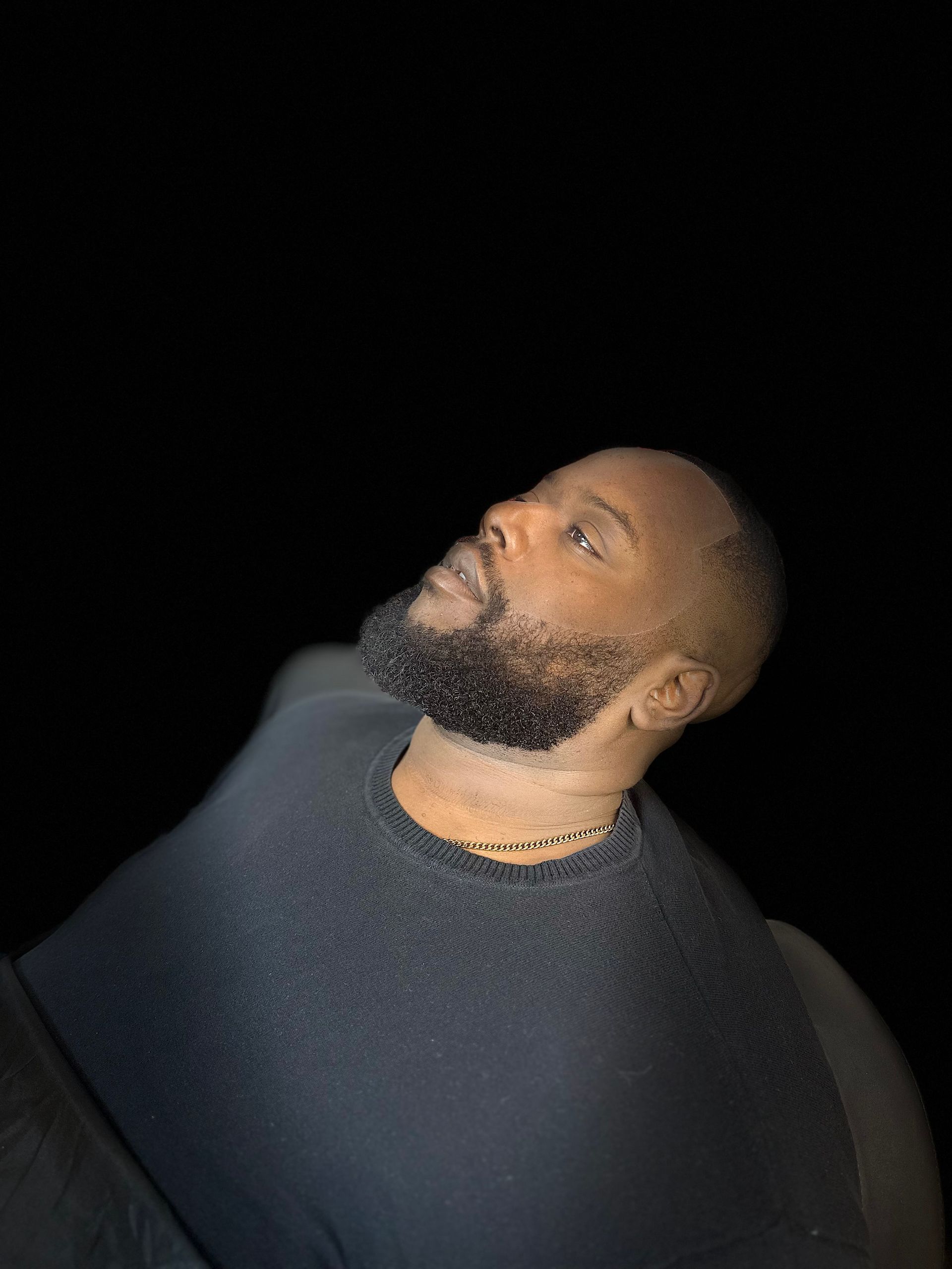 A man with a short beard and dark sweater looks upward against a black background.