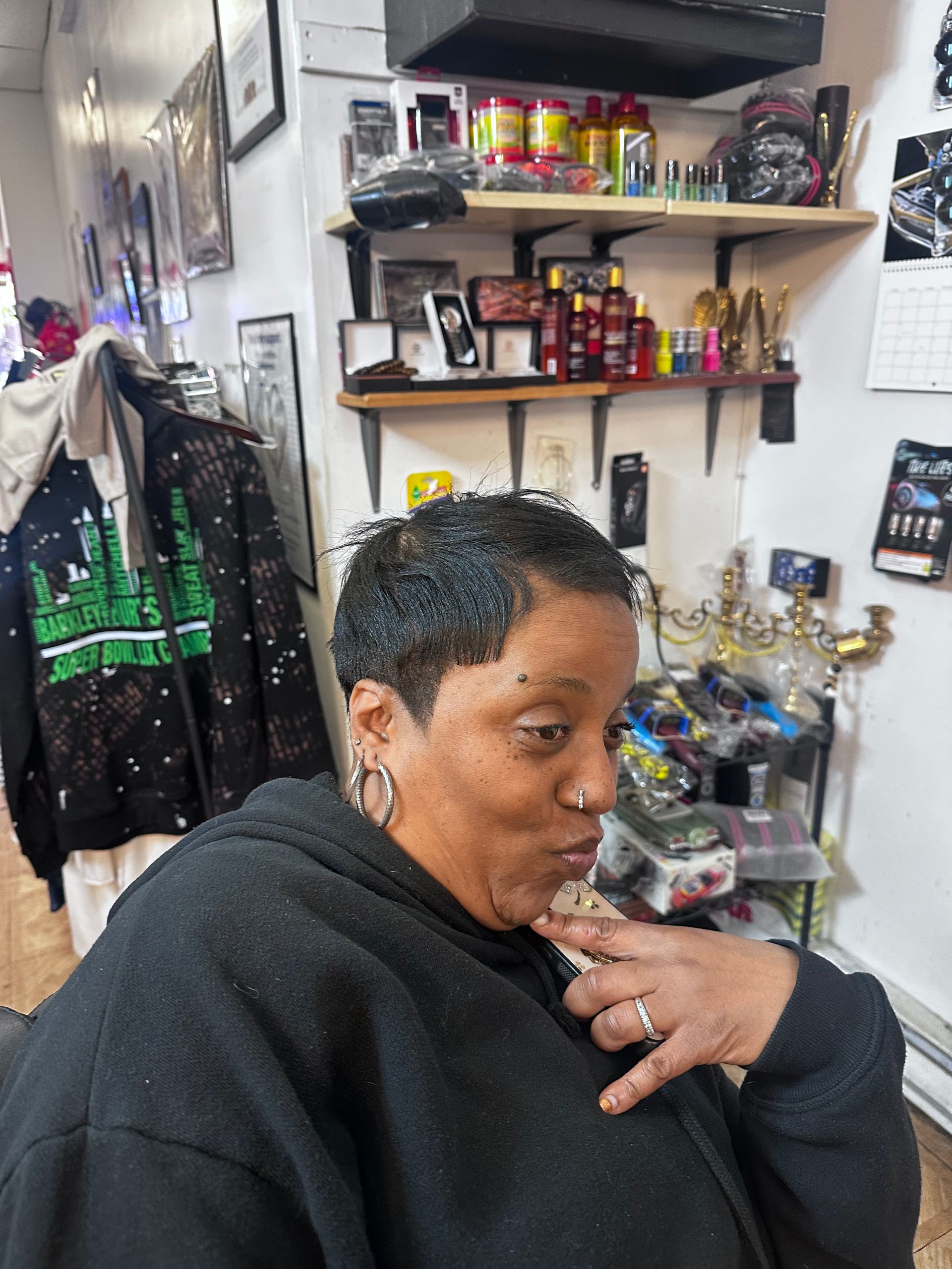 A person with short, dark, textured hair poses in a room filled with various bottles and tools on shelves.