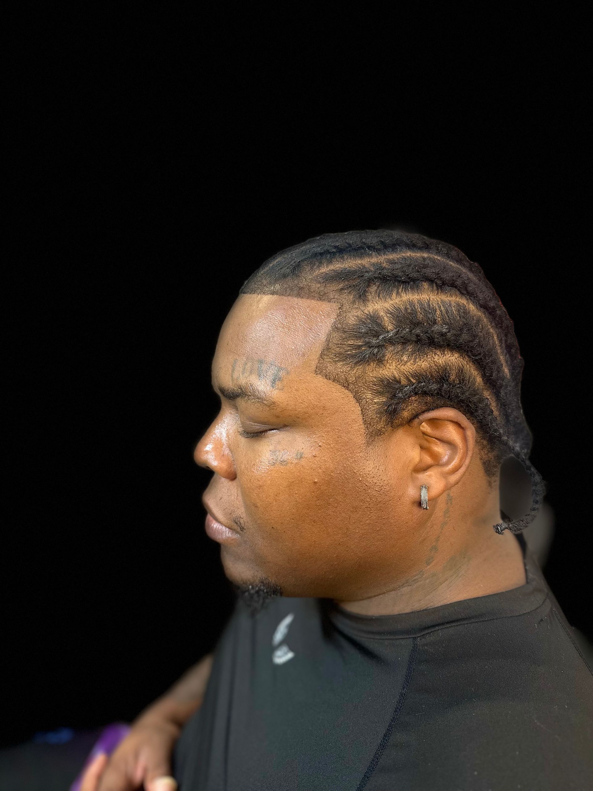 Profile view of a person with cornrowed hair and a clean-shaved hairline against a black background.