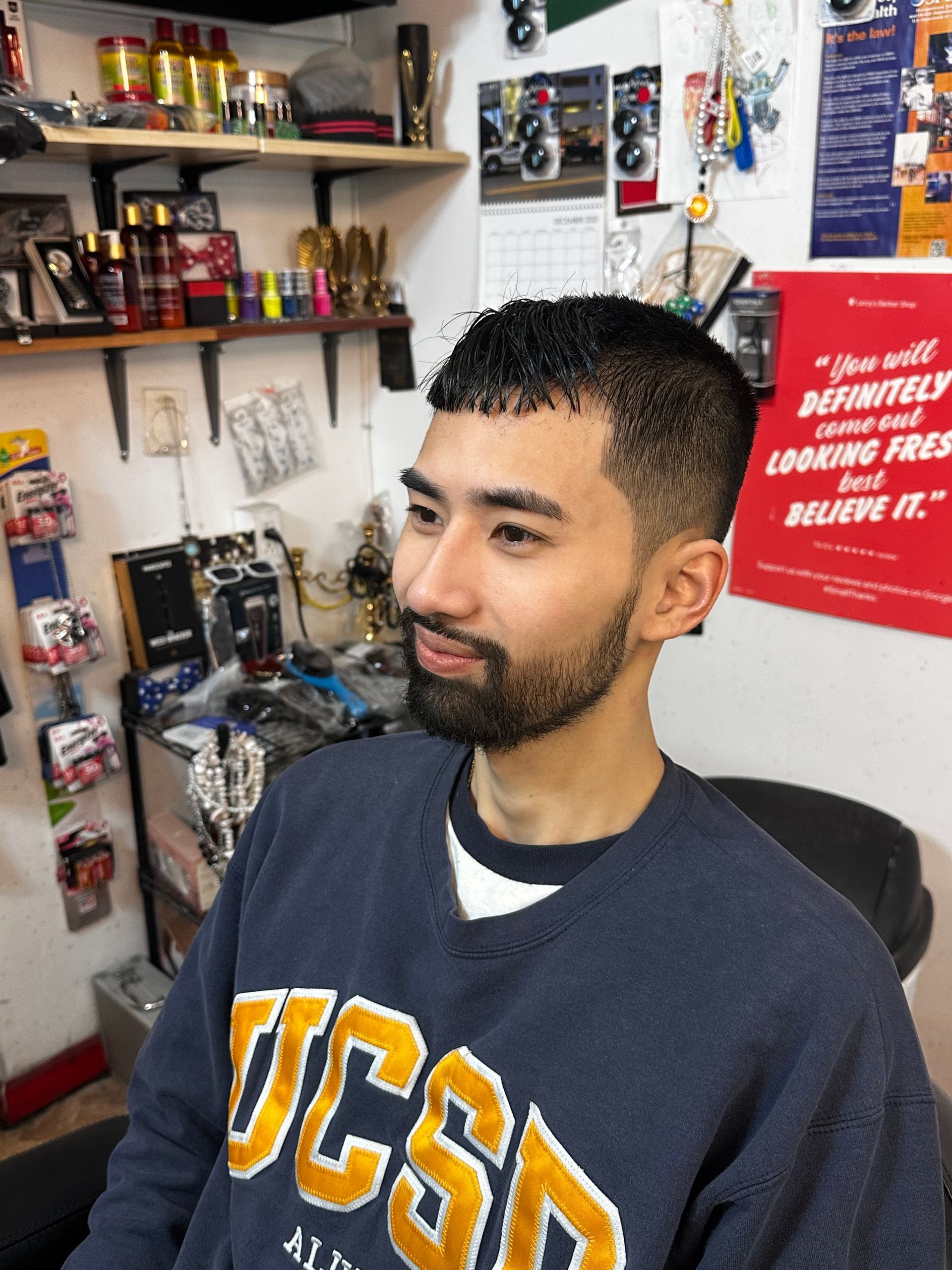 A person with a short, faded haircut and a groomed beard sits in a barbershop, wearing a blue UCSD sweatshirt.