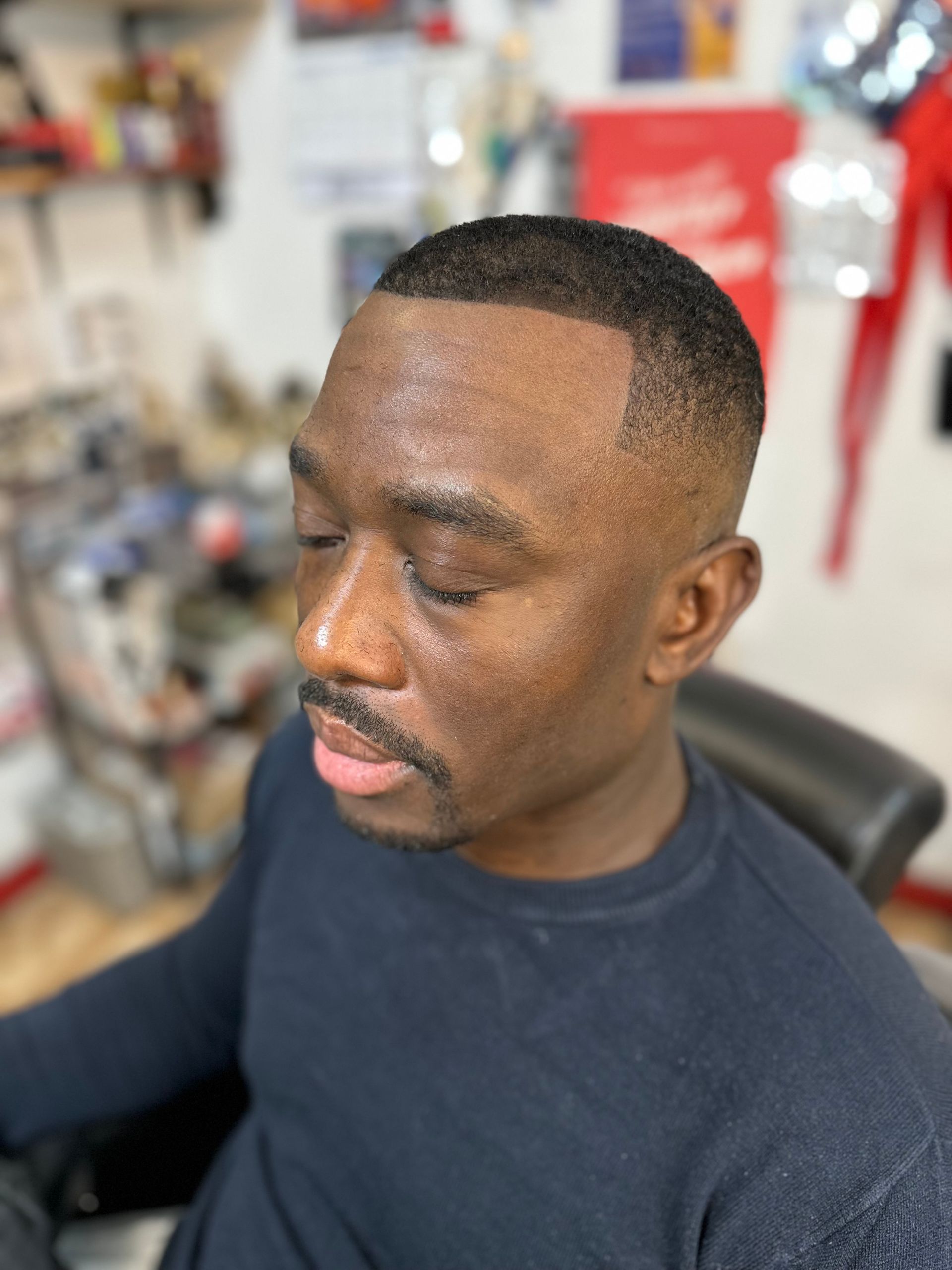 A man with a short, faded haircut and a thin mustache sits in a barbershop chair, looking down with closed eyes.