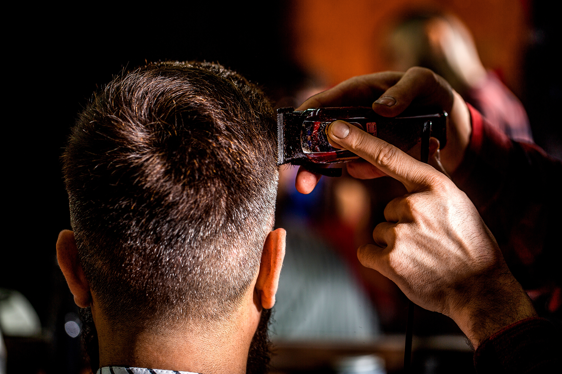 A barber using clippers to trim the hair on the back of a person's head in a dimly lit setting.
