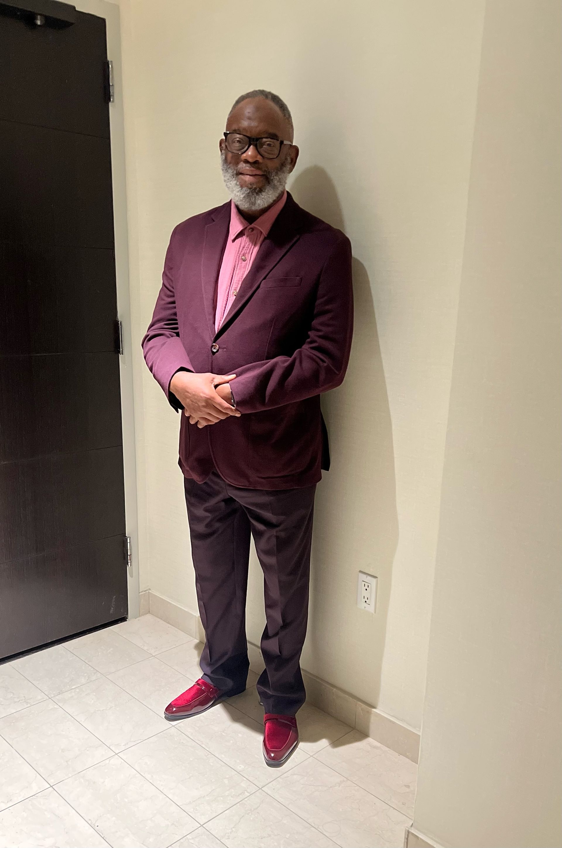 Two men standing side-by-side in a narrow barbershop, one in a black shirt and the other in a blue Philadelphia 76ers top.